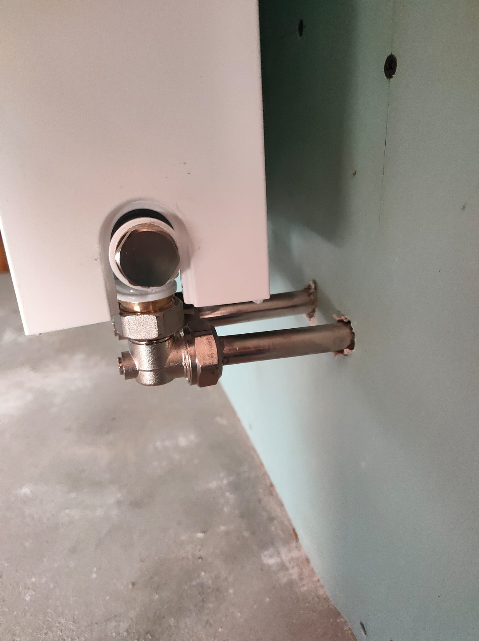 Szczegółowe ujęcie instalacji rur podłączeniowych do białego urządzenia grzewczego, z widocznymi mosiężnymi złączkami i fragmentem ściany z gipsu.