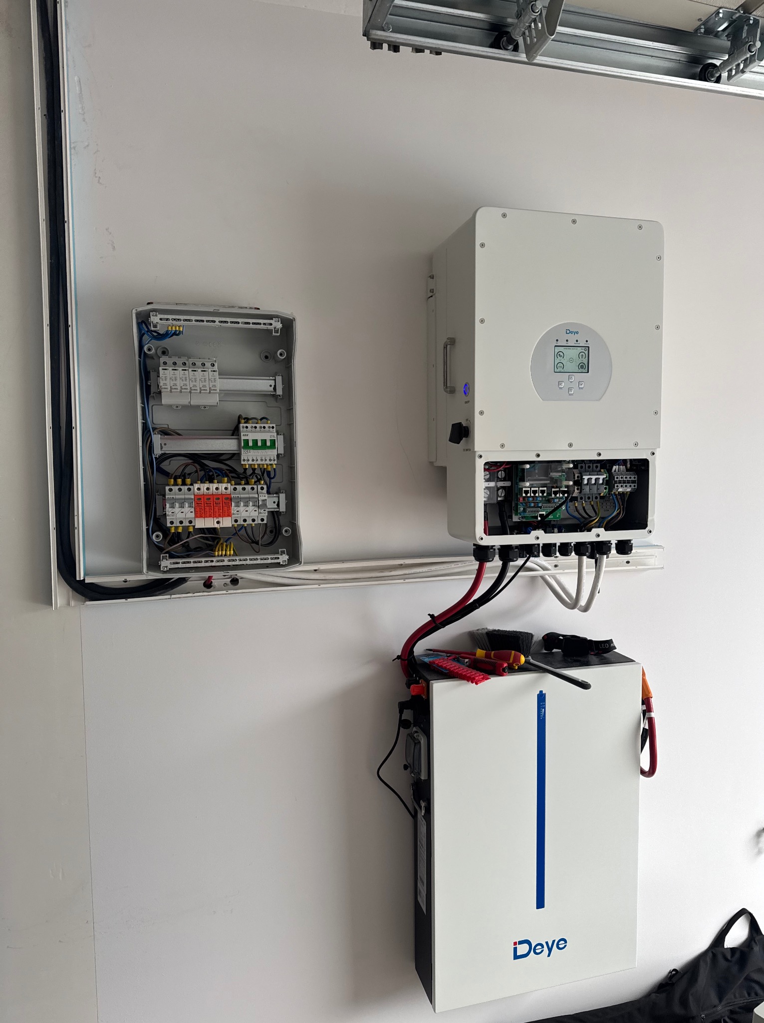 Rozdzielnica elektryczna, inwerter z wyświetlaczem i akumulator energii z logo Deye zamontowane na białej ścianie, widoczne kable i narzędzia