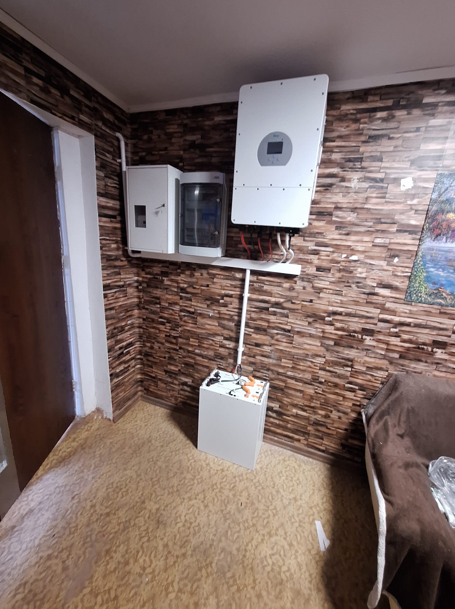 Wnętrze pomieszczenia z zamontowaną instalacją fotowoltaiczną: biały inwerter na ścianie wyłożonej panelami imitującymi cegłę, obok skrzynka rozdzielcza i akumulator, widoczne przewody i rury...