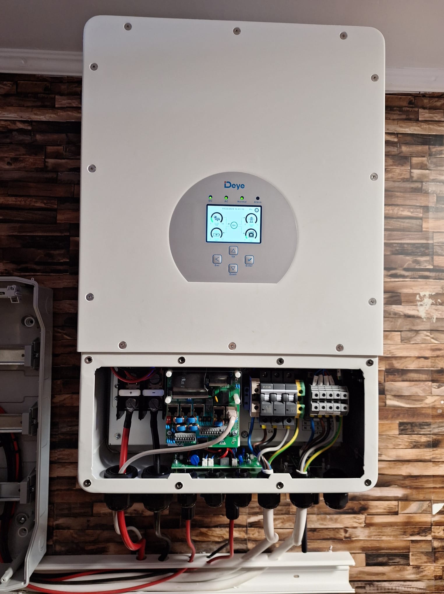 Otwarta obudowa falownika Deye z widocznymi elementami elektroniki wewnętrznej i wyświetlaczem LCD prezentującym parametry pracy, zamontowana na tle ściany z imitacji cegły.