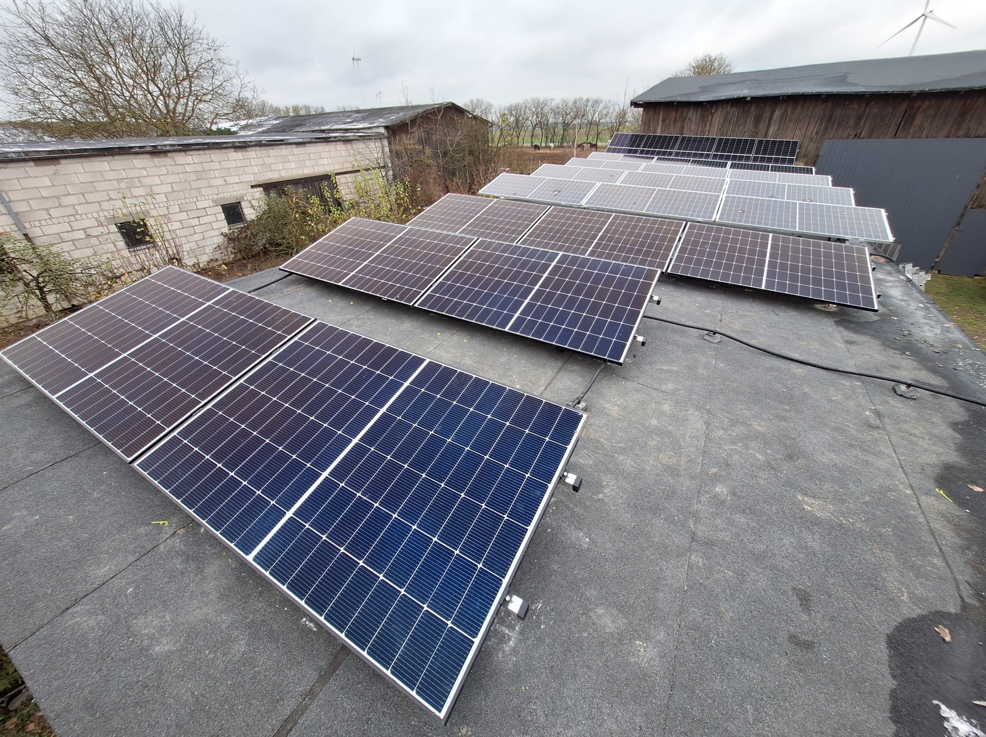 Rząd paneli słonecznych na płaskim dachu budynku gospodarczego, widoczne elementy montażowe i przewody, w tle zabudowania gospodarcze i drzewa bez liści, pochmurne niebo.
