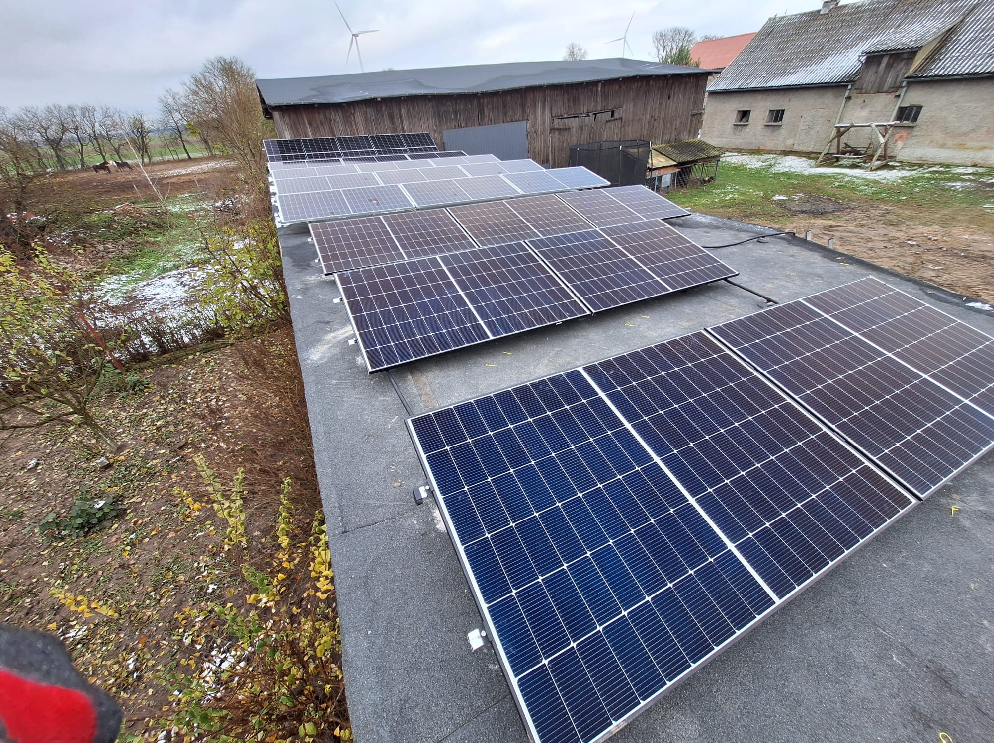 Rząd paneli słonecznych na płaskim dachu budynku gospodarczego, w tle farma w wietrzny, pochmurny dzień.
