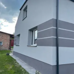 Świeżo wykończona elewacja domu jednorodzinnego z szarego tynku z poziomymi pasami w ciemniejszym odcieniu szarości, widoczny fragment rynny i chodnika wokół budynku.