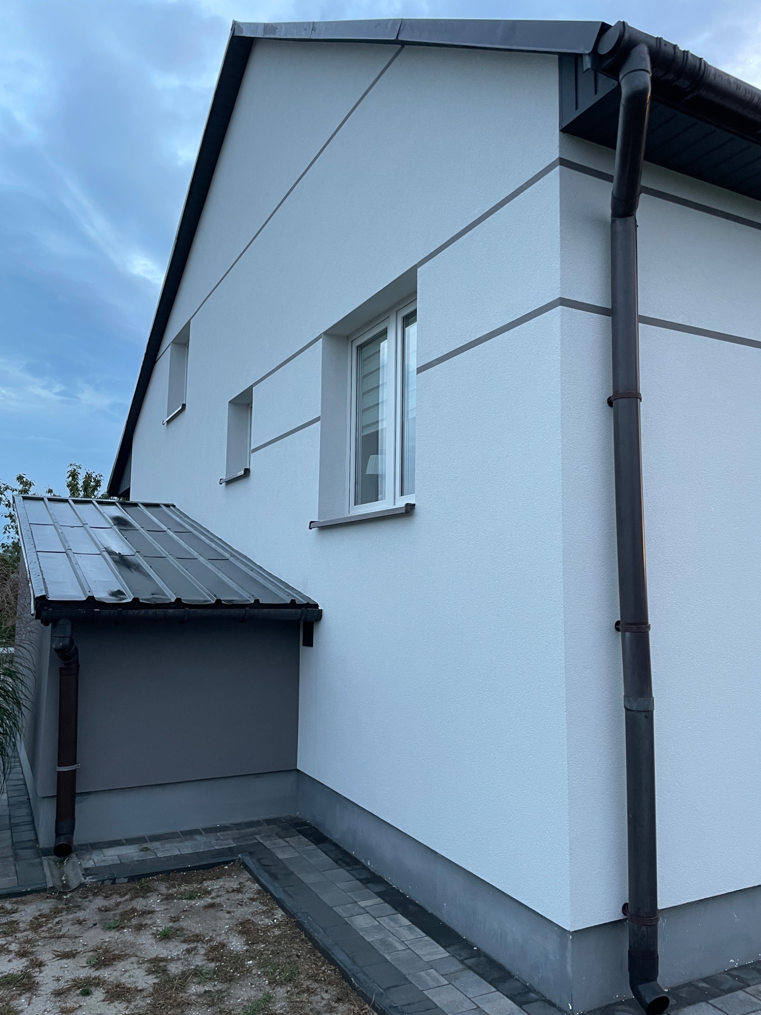 Nowoczesny dom jednorodzinny z białą elewacją, ciemnymi akcentami w postaci pasów i rynien, oraz zadaszeniem nad wejściem, widok z boku.