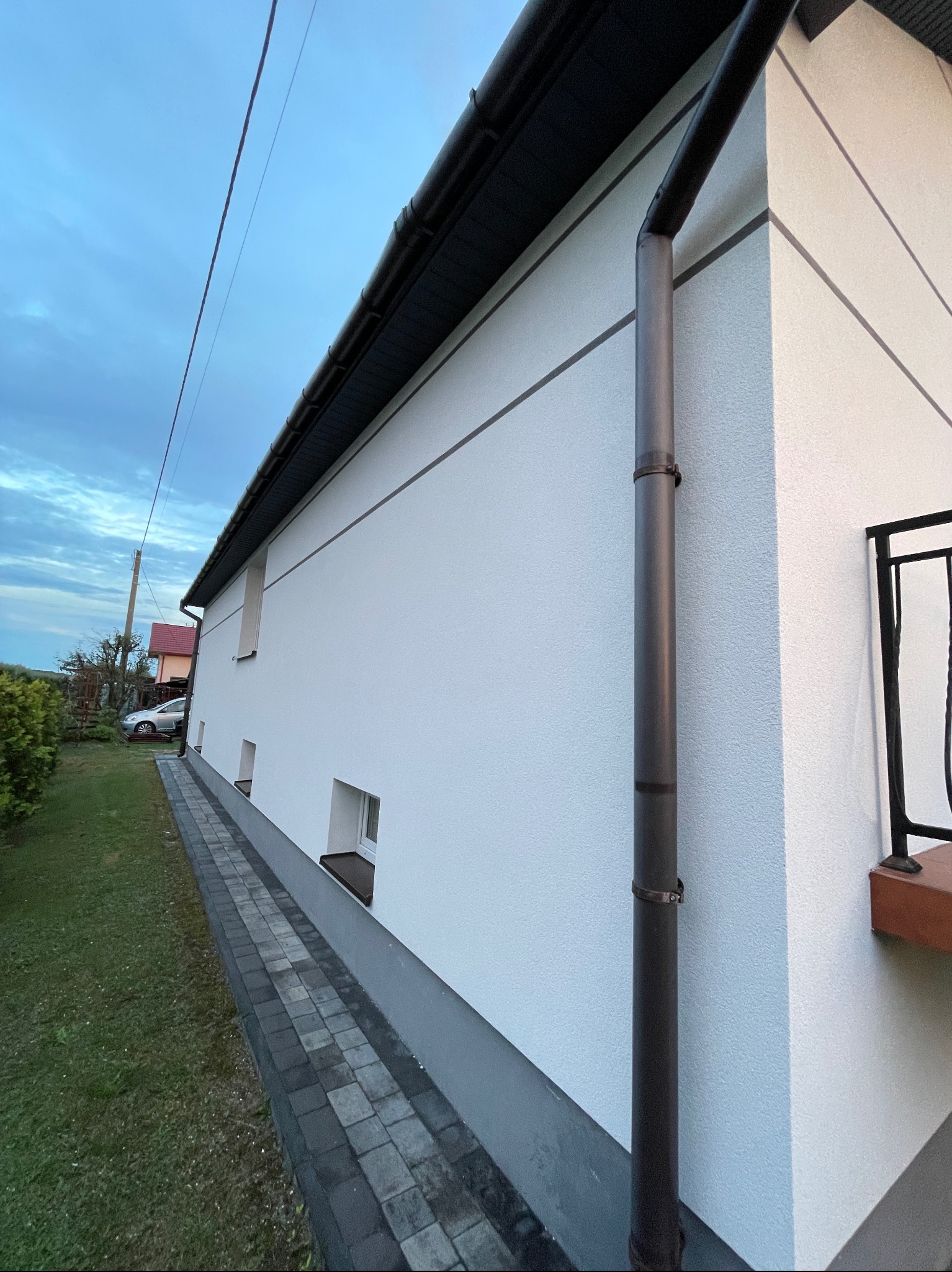Boczny widok otynkowanego na biało domu z brązową rynną i obróbkami blacharskimi, widoczny fragment chodnika z kostki brukowej, trawnik i fragment balkonu.