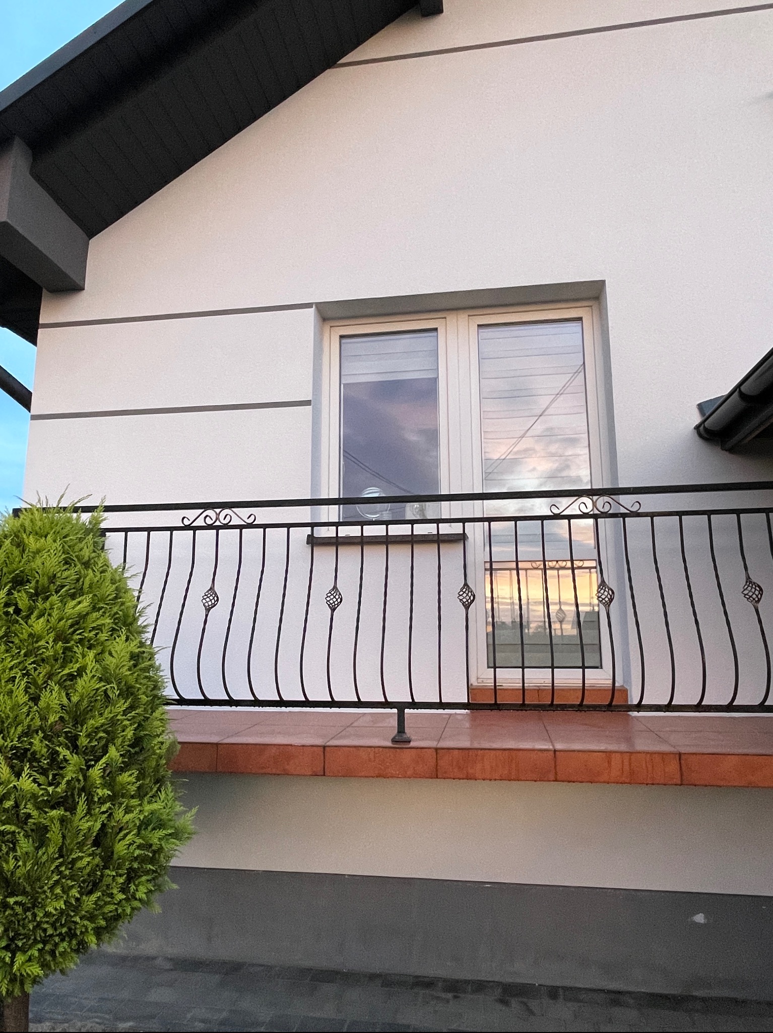 Elewacja domu jednorodzinnego z balkonem z dekoracyjną, kutą balustradą i widocznym odbiciem zachodu słońca w oknach.
