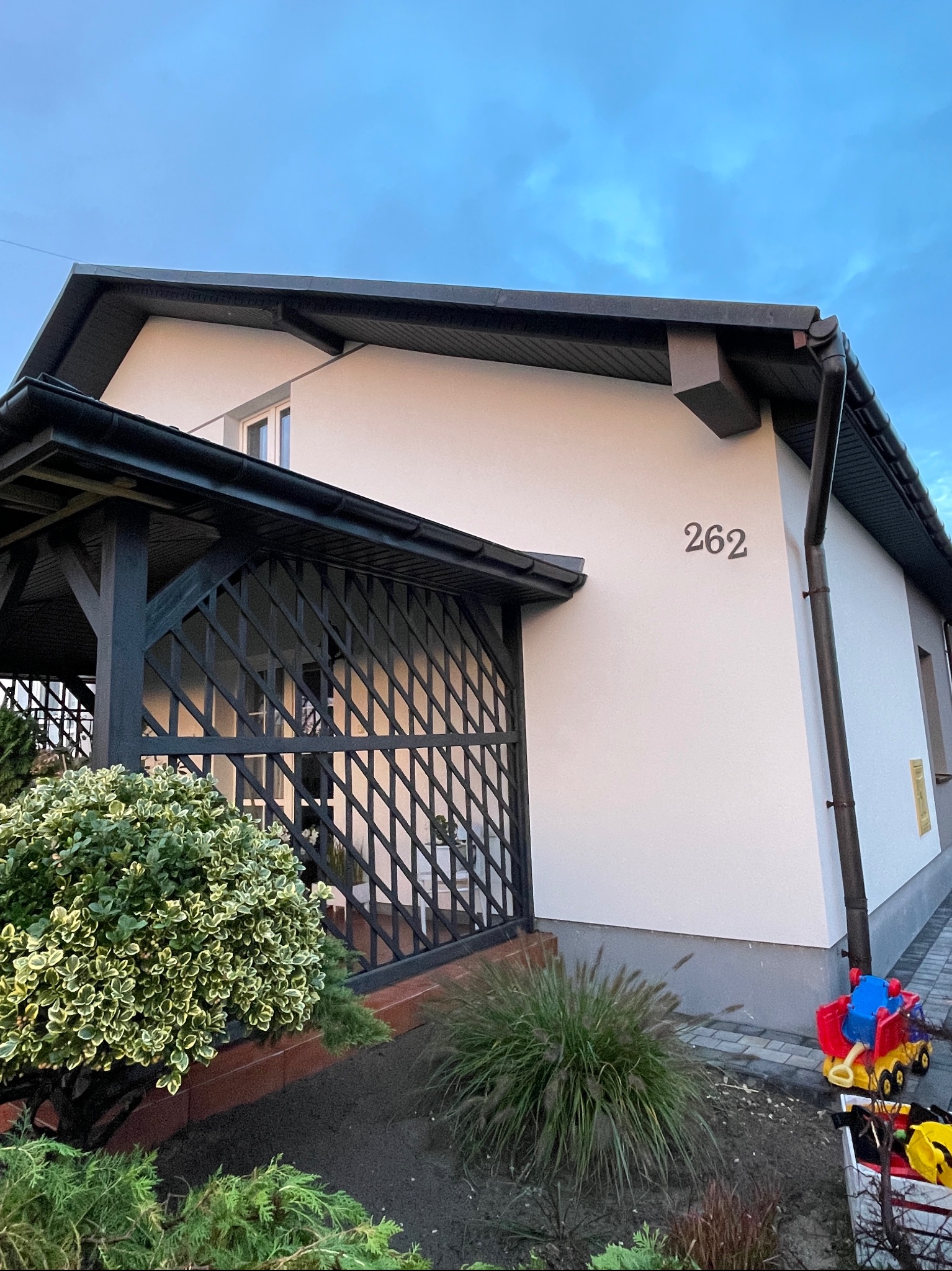 Ocieplony dom jednorodzinny z numerem 262, z czarną pergolą i ozdobnymi krzewami na rabatach, zabawki dziecięce przy rynnie.