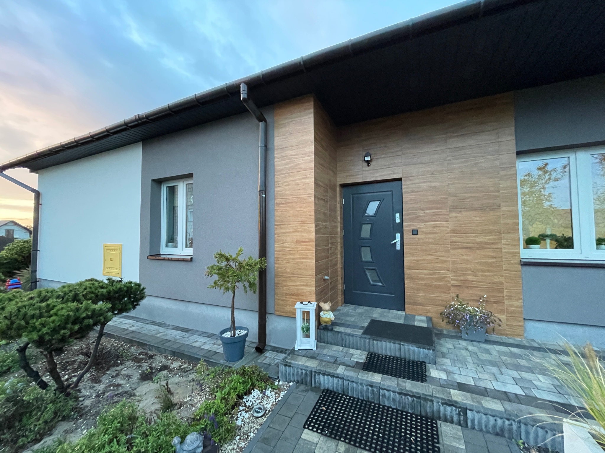 Nowoczesny dom jednorodzinny z elewacją w odcieniach szarości i drewna, widok na front z wejściem, schody wyłożone kostką, dekoracyjny lampion przy drzwiach, małe drzewko w donicy.