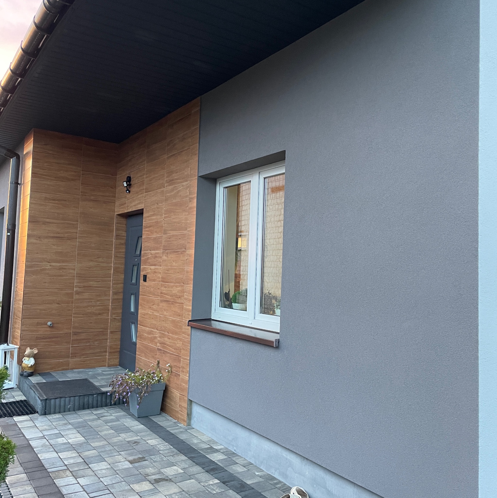 Elewacja domu jednorodzinnego z szarą strukturą tynku, brązową okładziną imitującą drewno przy wejściu i białymi oknami, widok z zewnątrz na kostkę brukową i donicę z kwiatami.