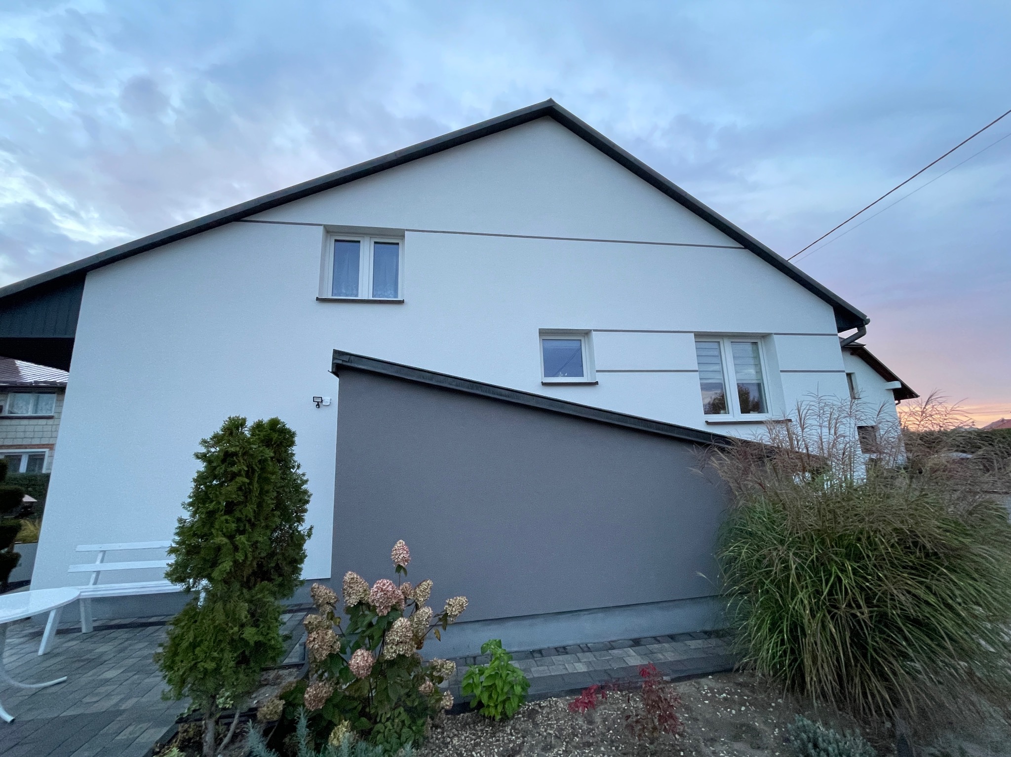 Dwukondygnacyjny dom z białą elewacją, szarą dobudówką i ciemnym dachem, widoczny z perspektywy ogrodu z ozdobnymi trawami i tujami.