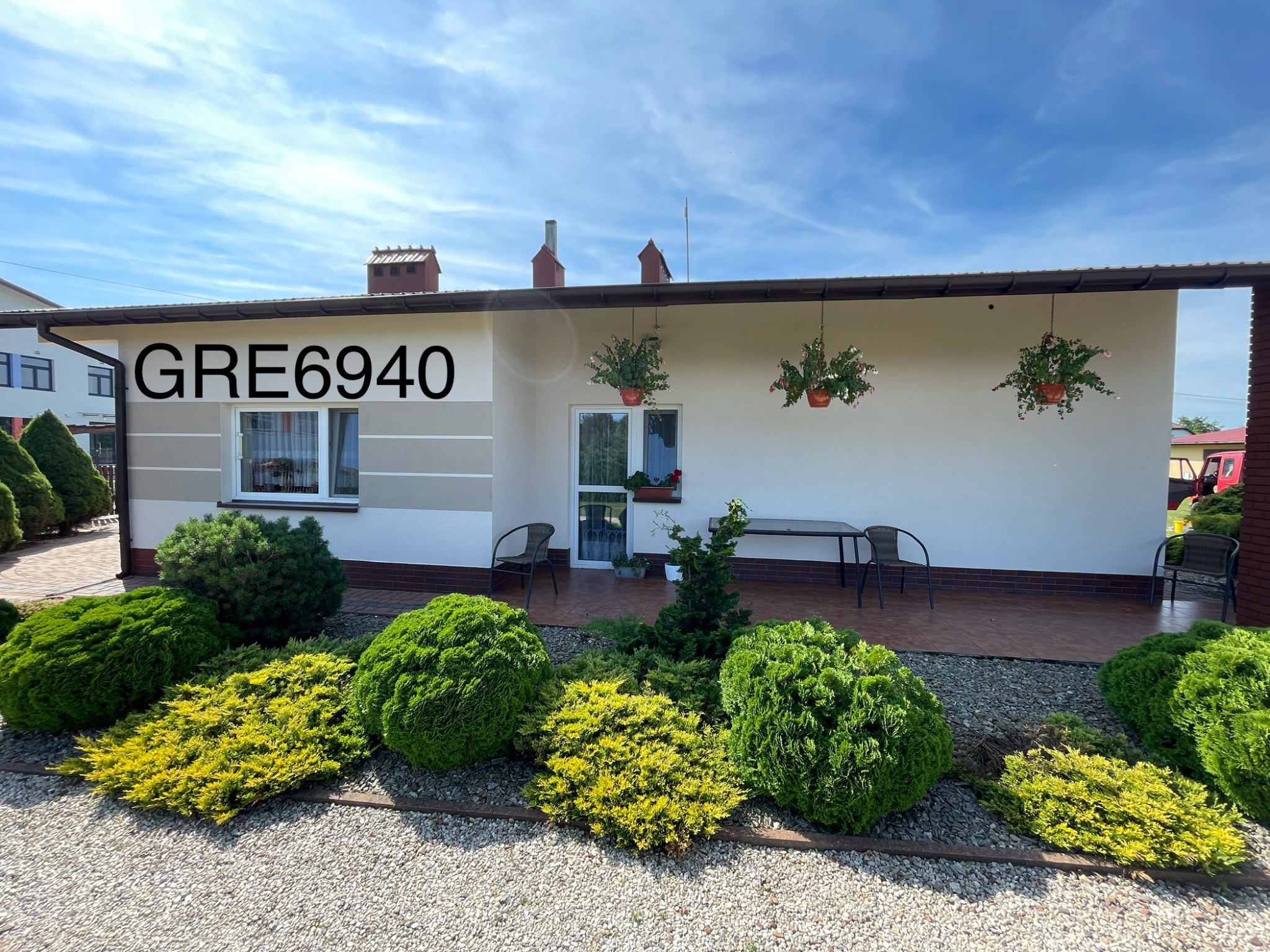 Parterowy dom jednorodzinny z białymi ścianami i brązowym dachem, z zadbanym ogrodem z krzewami iglastymi i kwiatami w wiszących donicach, widoczny numer domu na ścianie.