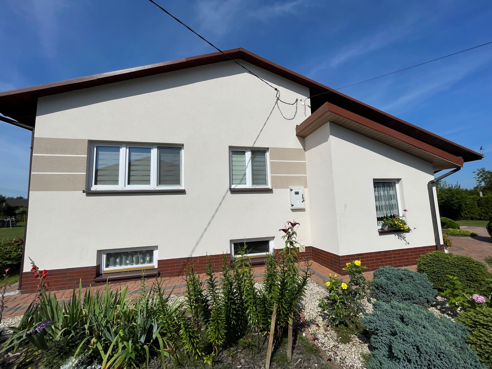 Jasny dom jednorodzinny z kremową elewacją, brązowym dachem i detalami z cegły klinkierowej, otoczony zielenią i kwiatami w słoneczny dzień.