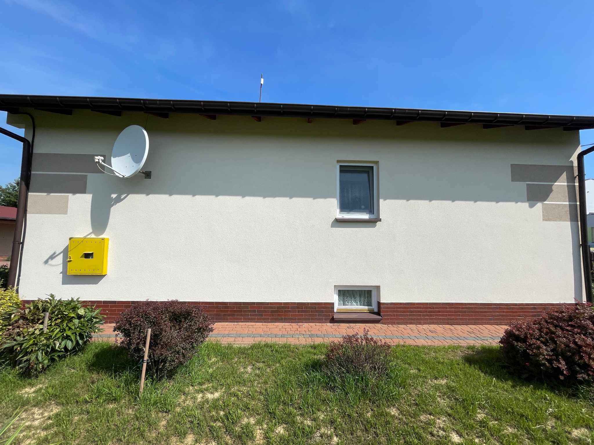 Fragment elewacji domu jednorodzinnego z jasną elewacją, brązowym pasem z cegły klinkierowej u podstawy, białym oknem, anteną satelitarną i żółtą skrzynką gazową, otoczony zielenią.