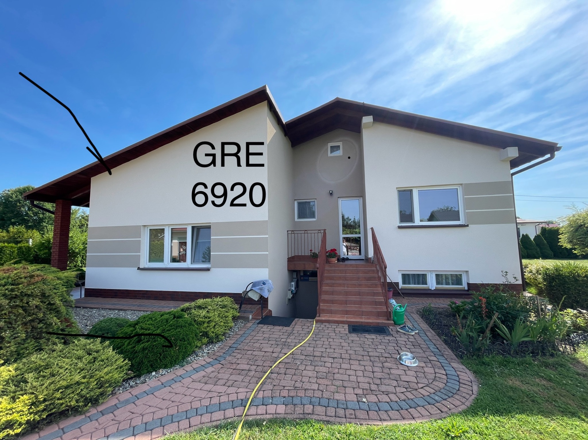 Dwukondygnacyjny dom jednorodzinny z jasną elewacją i brązowymi akcentami, widoczny z poziomu ogrodu z kostki brukowej, z numerem domu 'GRE 6920' widocznym na ścianie.