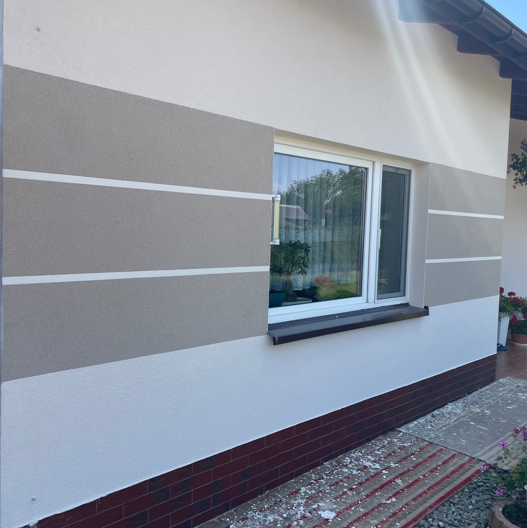 Elewacja domu po ociepleniu z dekoracyjnymi szarymi pasami i białymi liniami oddzielającymi, widoczne okno z roślinami na parapecie, fragment chodnika z resztkami styropianu.