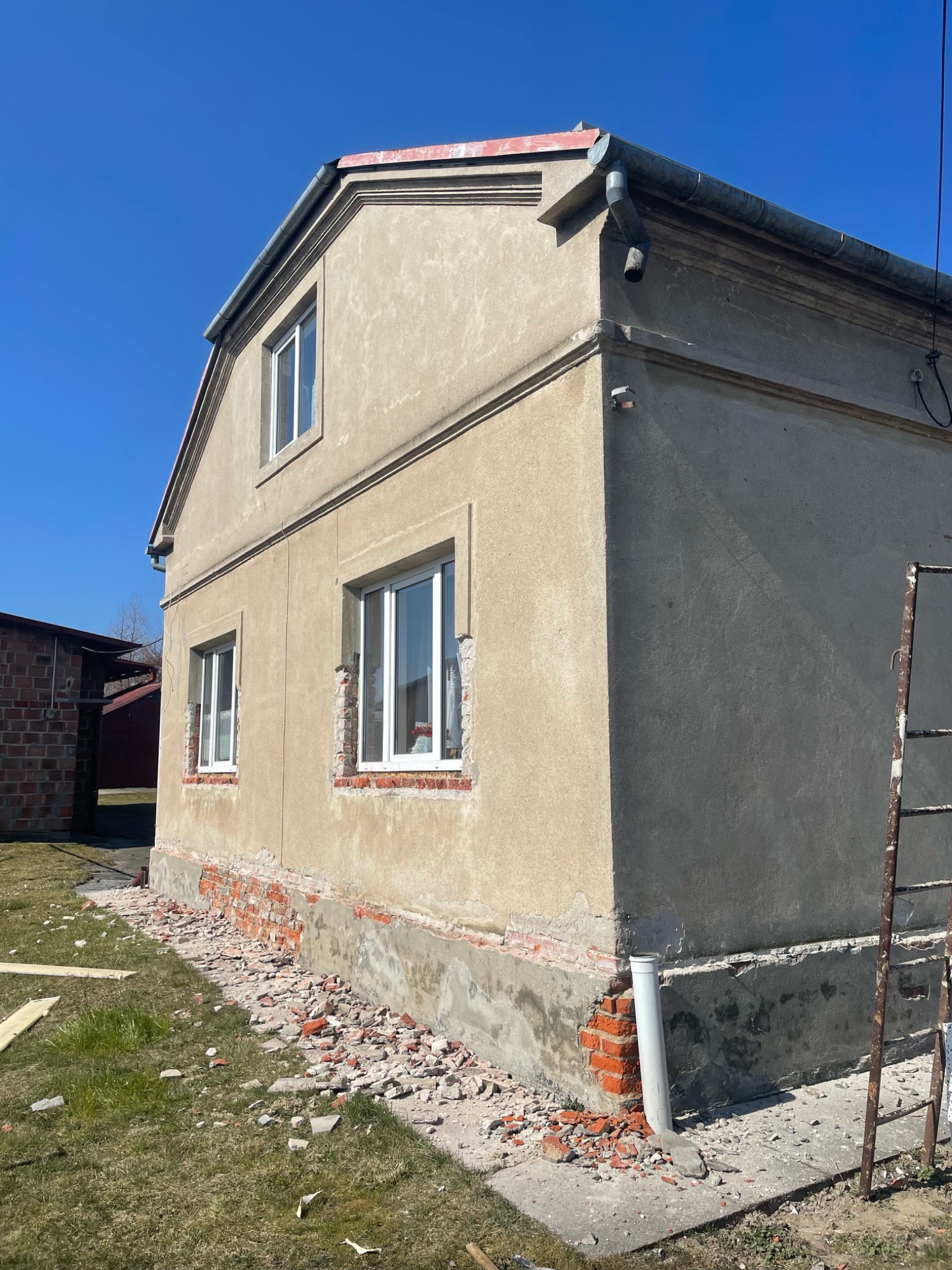 Dom z odsłoniętą cegłą wokół okien i fundamentów, przygotowywany do prac elewacyjnych, z widocznymi resztkami gruzu i drabina w tle