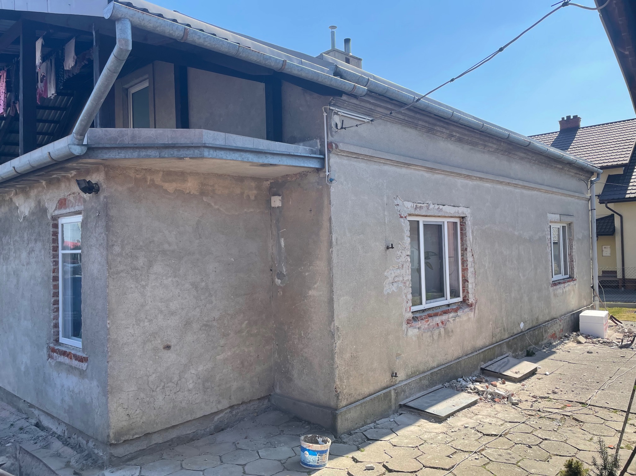 Dom jednorodzinny z widocznymi śladami prac remontowych elewacji, z odsłoniętą cegłą wokół okien, pozostałościami materiałów budowlanych na podwórku i wiaderkiem z zaprawą przy ścianie.