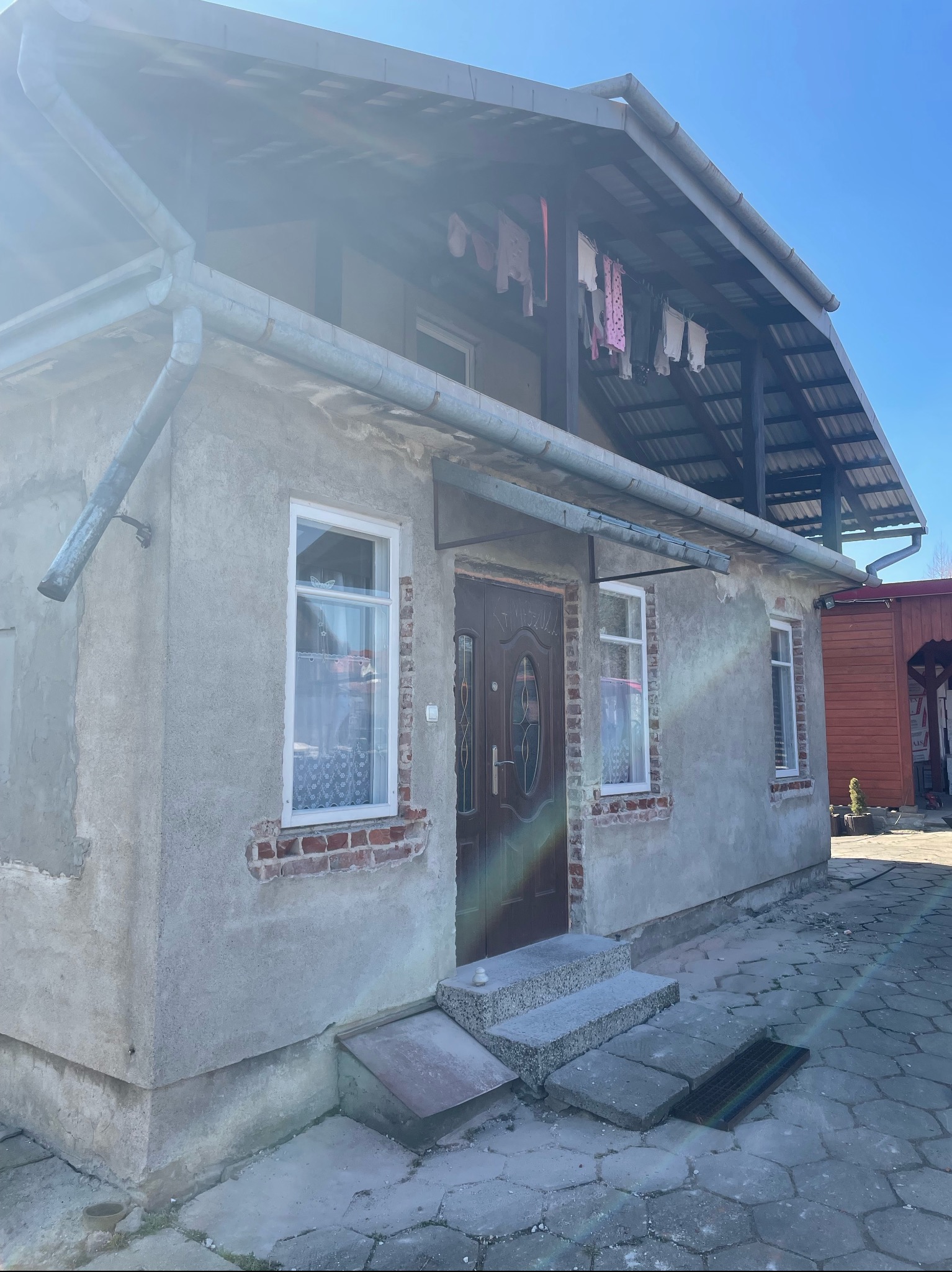 Dom jednorodzinny przed remontem elewacji, widoczne stare okna, zniszczony tynk i fragmenty odsłoniętej cegły, z elementami prania suszącego się na balkonie.