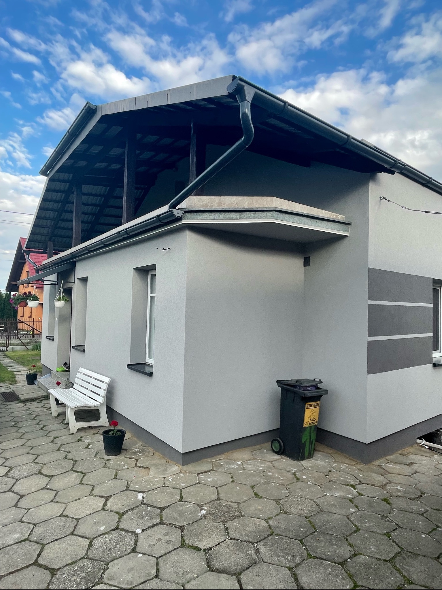 Szary dom jednorodzinny z ocieploną elewacją, widoczny z boku, z ciemnymi pasami na ścianie, białą ławeczką i koszem na śmieci obok, na brukowanym podwórku.