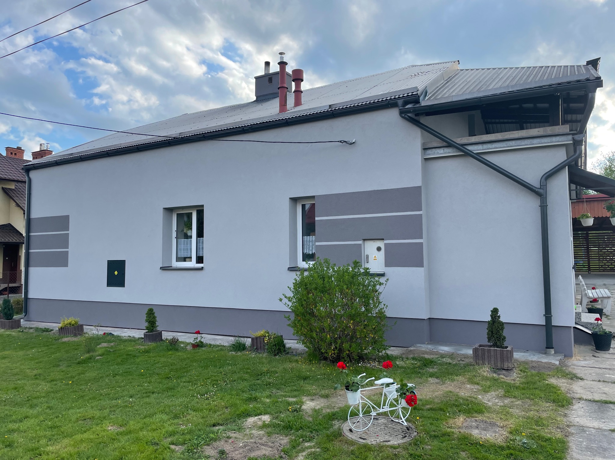 Odnowiona elewacja domu jednorodzinnego w odcieniach szarości z dekoracyjnymi pasami, widoczne czerwone kominy, ozdobny kwietnik w kształcie roweru i zadbany trawnik.