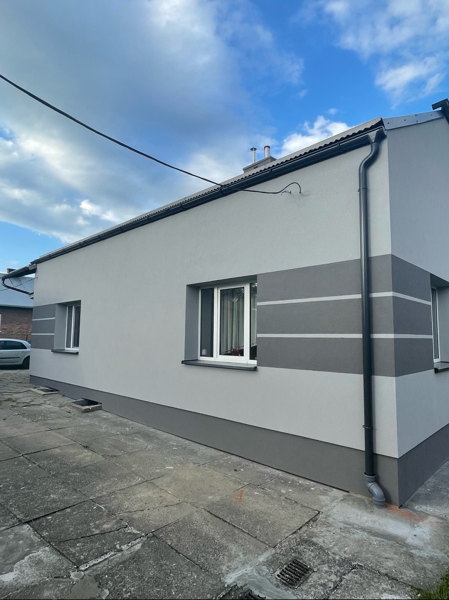 Świeżo otynkowany dom jednorodzinny w odcieniach szarości, z ciemniejszym pasem na dole i trzema poziomymi pasami na elewacji, widoczne okna z białymi ramami i czarne rynny.