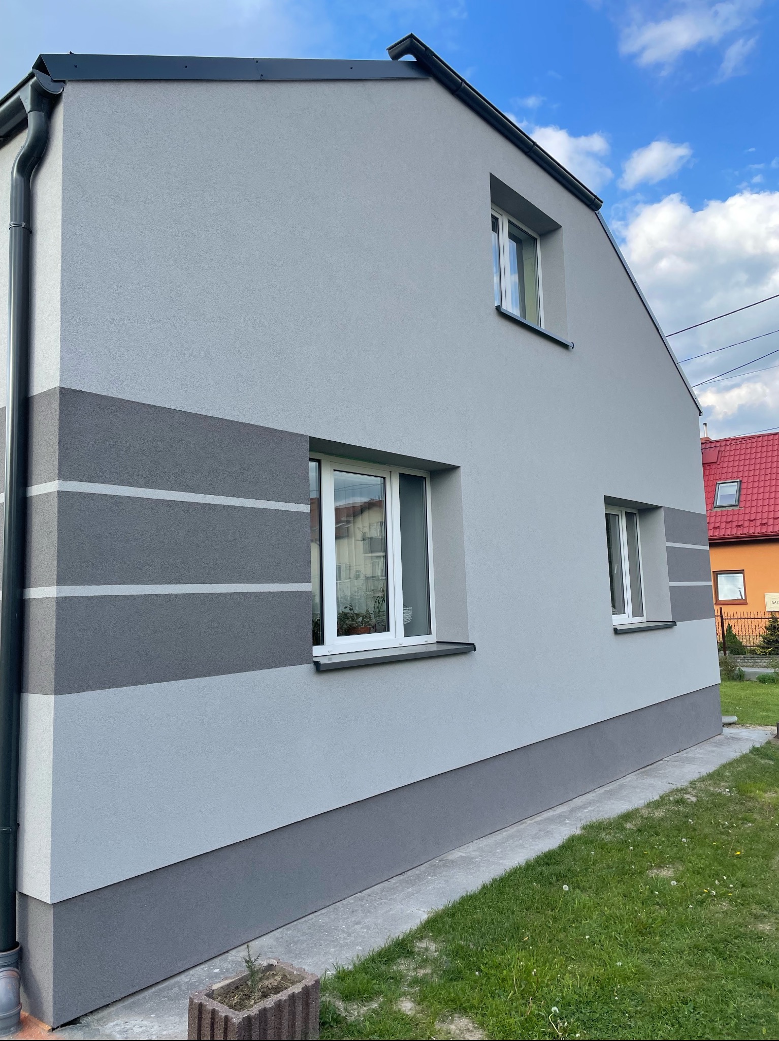 Świeżo otynkowana ściana domu jednorodzinnego w odcieniach szarości z poziomymi pasami, widoczne okna z białymi ramami, rynna i fragment trawnika.