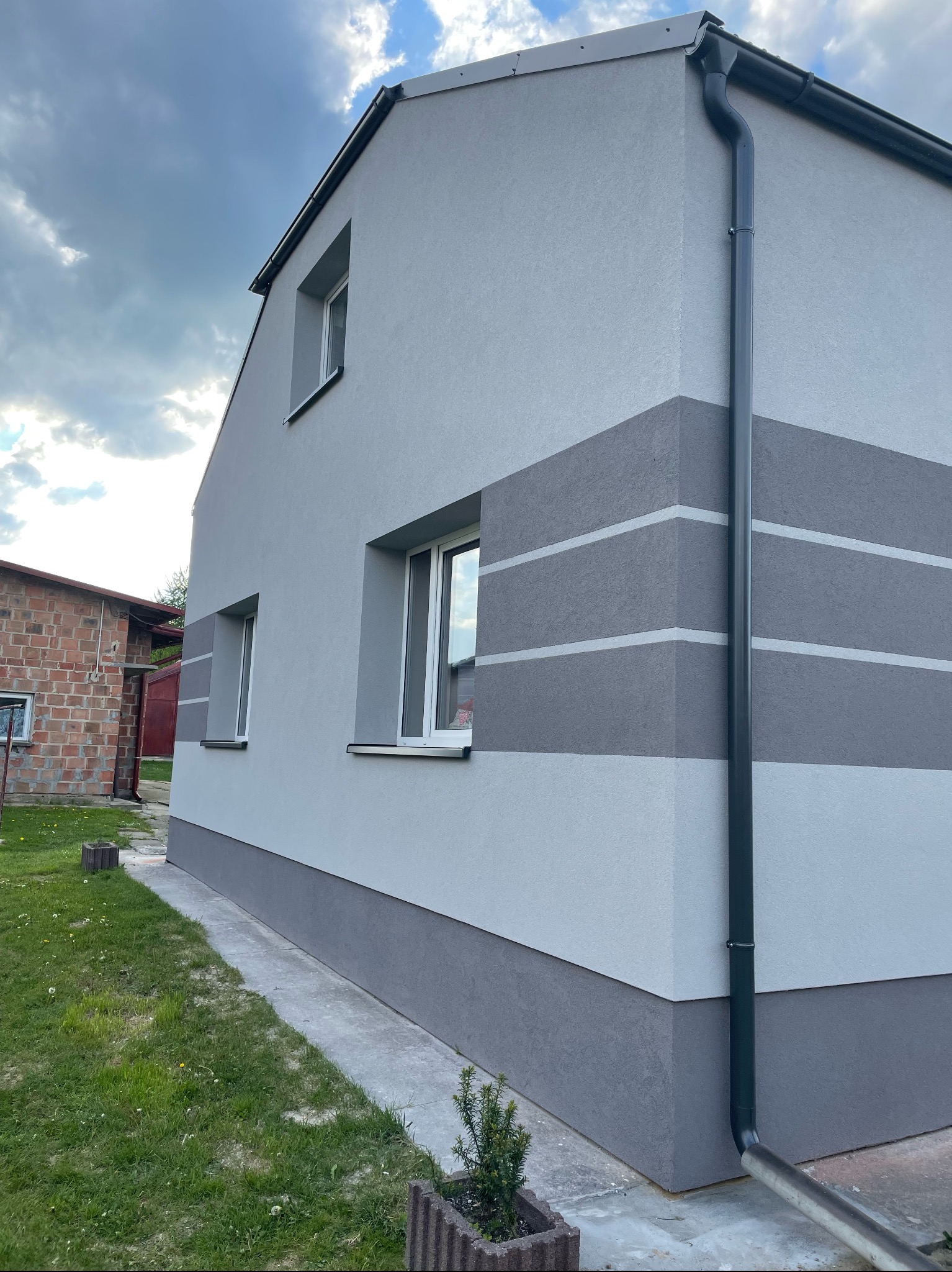 Świeżo wykończona elewacja domu jednorodzinnego z szarego tynku z poziomymi pasami w ciemniejszym odcieniu szarości, widoczny fragment rynny i chodnika wokół budynku.