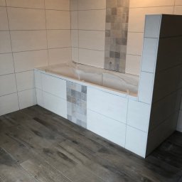 Tomski - Wykończona łazienka z drewnopodobną podłogą, białą zabudową wanny z szarymi akcentami mozaiki i świeżo położonymi białymi płytkami na ścianach.