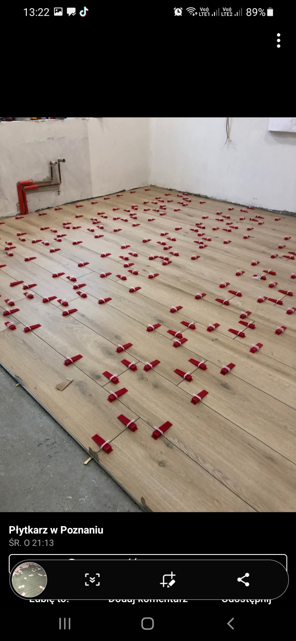 Ułożone panele podłogowe imitujące drewno, z czerwonymi klinami dystansowymi i białymi gumkami, na tle surowych ścian w trakcie prac wykończeniowych.