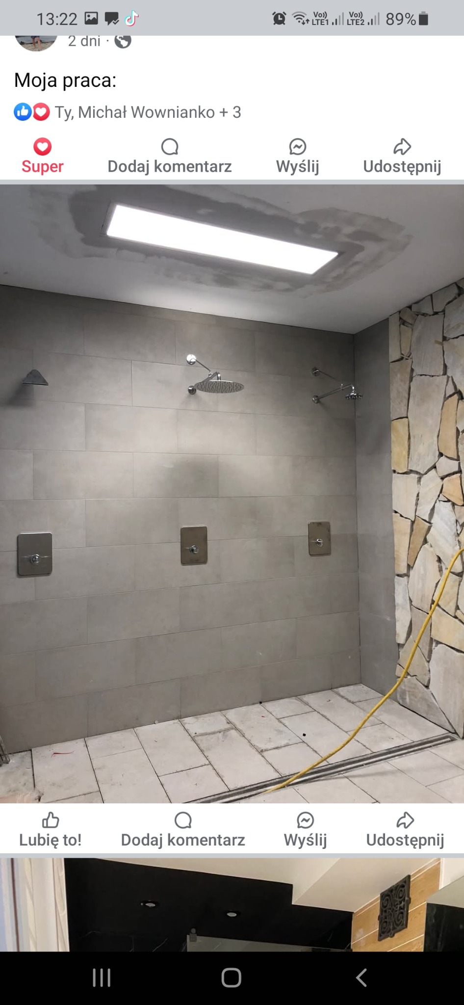 Nowoczesna łazienka w trakcie wykończenia, z szarymi płytkami ściennymi, kamienną ścianą akcentową i trzema prysznicami z kwadratowymi regulatorami, widoczna listwa odpływowa w podłodze i żółty...