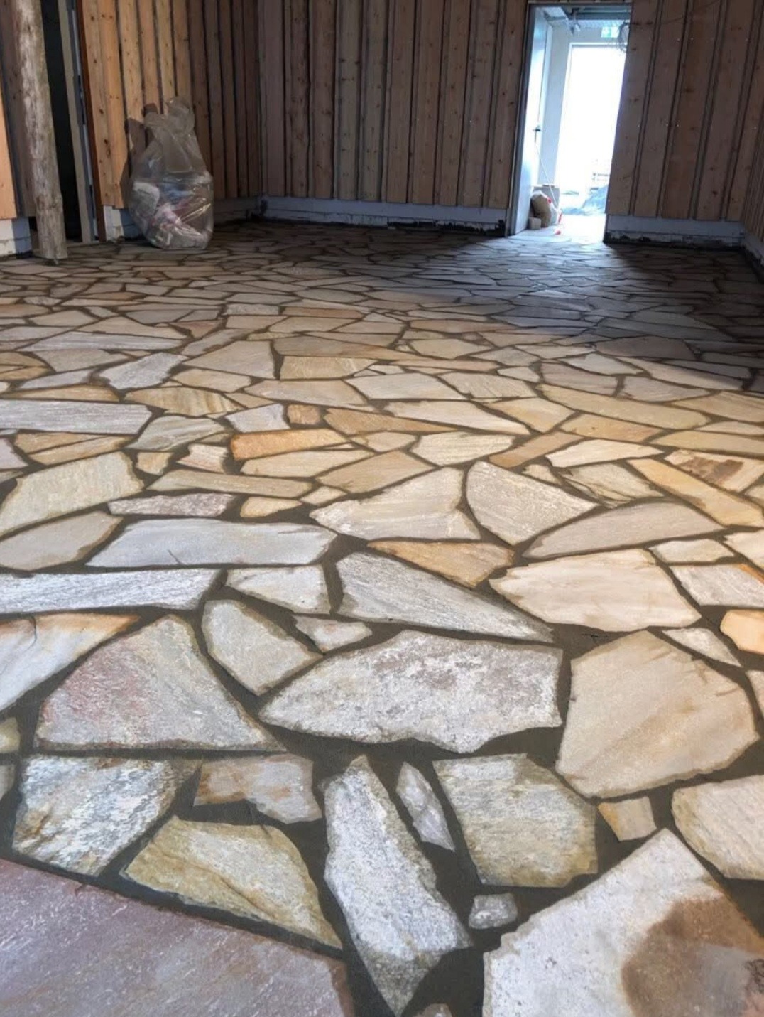 Podłoga wyłożona nieregularnymi płytami kamiennymi w pomieszczeniu z boazerią, widoczny worek z odpadami w rogu.