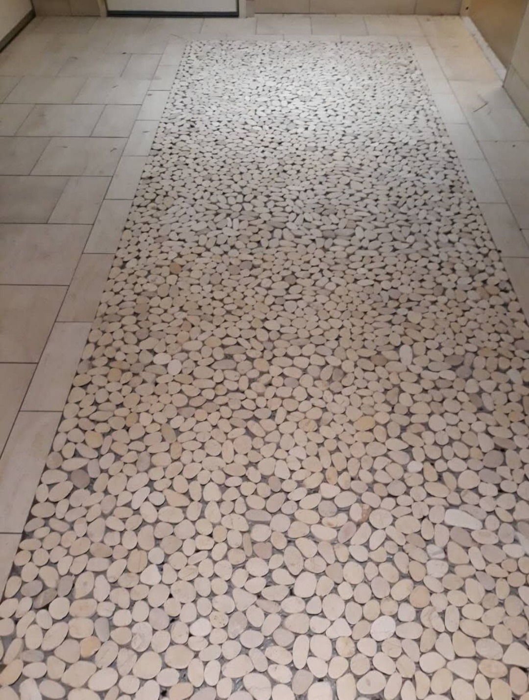 Dekoracyjna podłoga wyłożona jasnymi, owalnymi kamieniami rzecznymi, otoczona jasnymi płytkami podłogowymi, tworząca unikalny wzór przypominający ścieżkę.