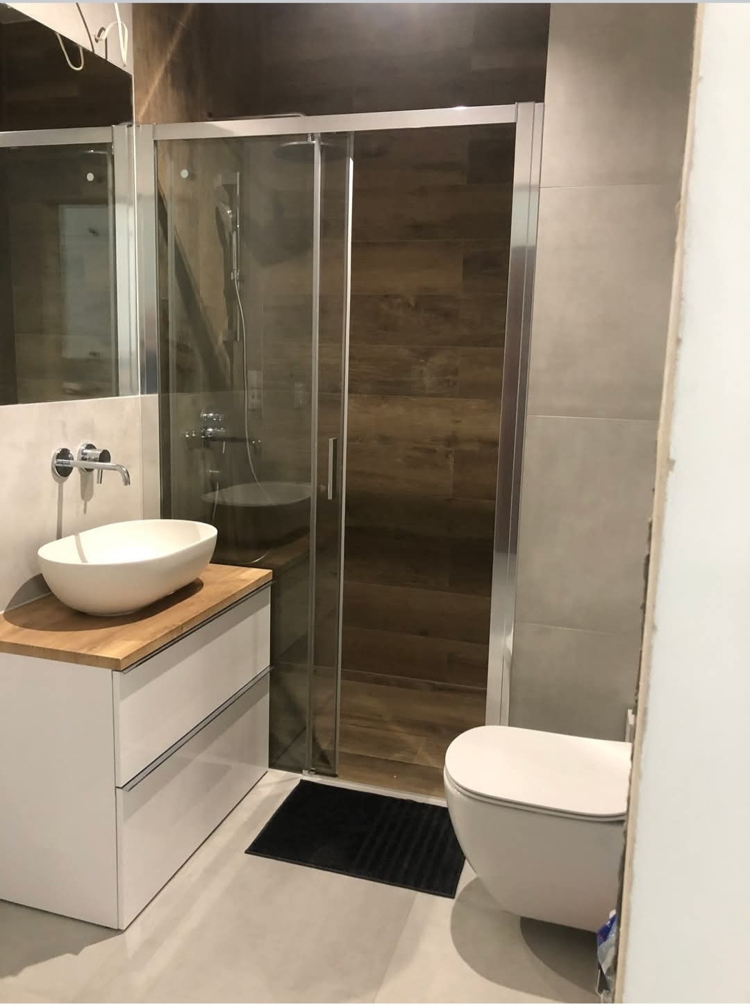 Nowoczesna łazienka z kabiną prysznicową z przesuwnymi drzwiami, umywalką nablatową na drewnianym blacie i wiszącą toaletą, wykończona płytkami imitującymi drewno i szarym gresem.