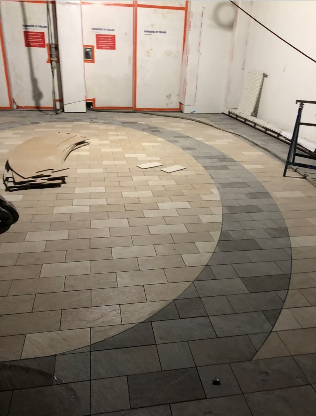 Układanie płytek podłogowych w okrągłym wzorze, z elementami w jasnych i ciemnych odcieniach szarości, widoczne przygotowane szablony i pozostałości po pracach w surowym wnętrzu.