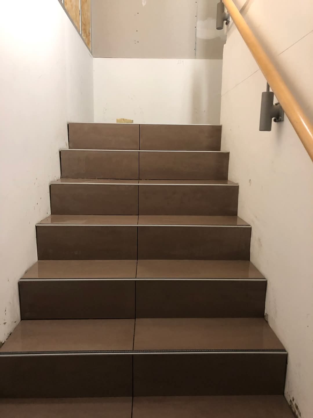 Schody wyłożone brązowymi płytkami z metalowymi listwami krawędziowymi, widoczna drewniana poręcz i fragment surowej ściany z płyt karton-gips.