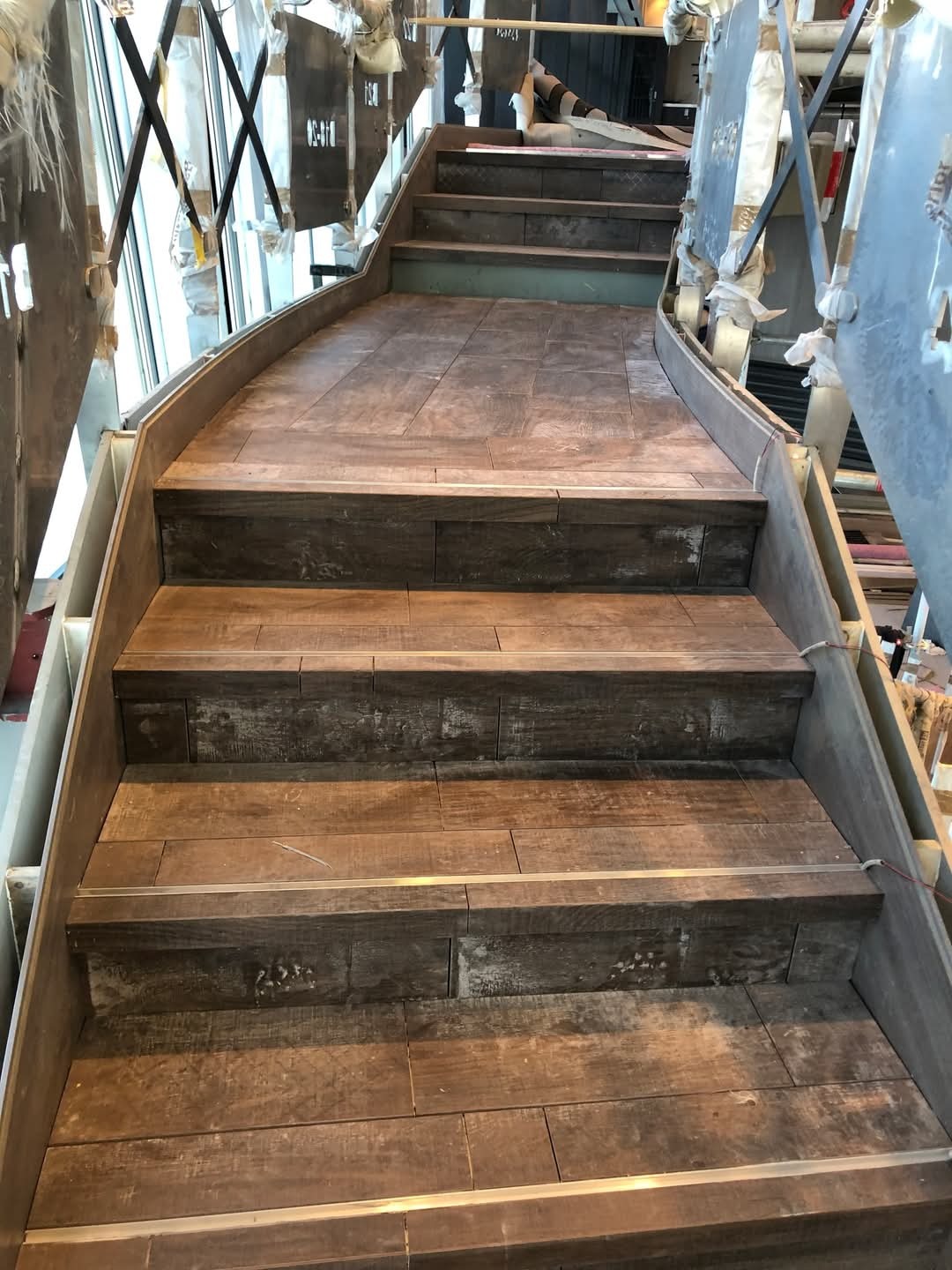 Schody z drewnopodobnych płytek w trakcie montażu, widoczne zabezpieczenia na elementach konstrukcyjnych oraz metalowe listwy na krawędziach stopni.