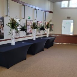 Efa Gryt Wynajem sprzętu plenerowego - Aranżacja we wnętrzu budynku z czterema czarnymi stołami nakrytymi obrusami, na których stoją wysokie, smukłe wazony z kompozycjami kwiatowymi i zielonymi gałązkami; w tle plakaty na ścianie...