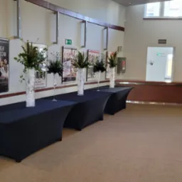 Aranżacja we wnętrzu budynku z czterema czarnymi stołami nakrytymi obrusami, na których stoją wysokie, smukłe wazony z kompozycjami kwiatowymi i zielonymi gałązkami; w tle plakaty na ścianie...