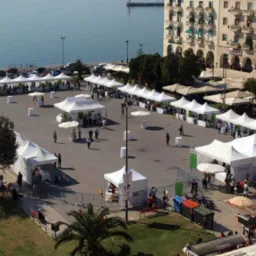 Widok z góry na plac miejski z rozstawionymi białymi namiotami, parasolami i stolikami, prawdopodobnie podczas festynu lub targów, z budynkami w tle i widokiem na morze.