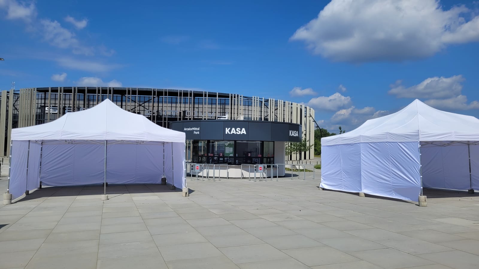 Dwa białe namioty plenerowe ustawione przed budynkiem z napisem 'KASA' w ArcelorMittal Park, błękitne niebo z chmurami w tle.