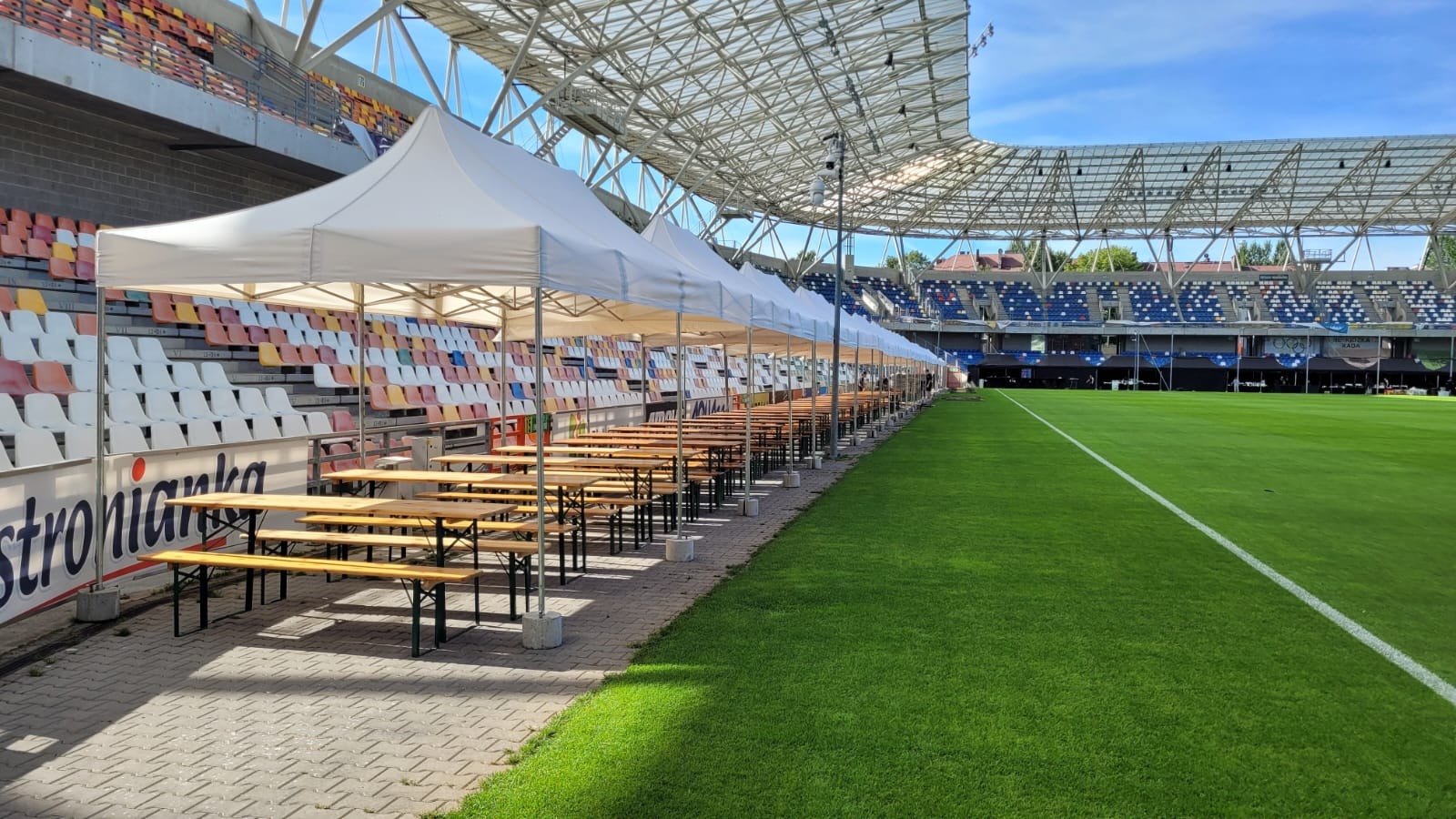 Ustawione rzędy białych namiotów z drewnianymi stołami i ławkami na stadionie, widok na zieloną murawę boiska i kolorowe trybuny.