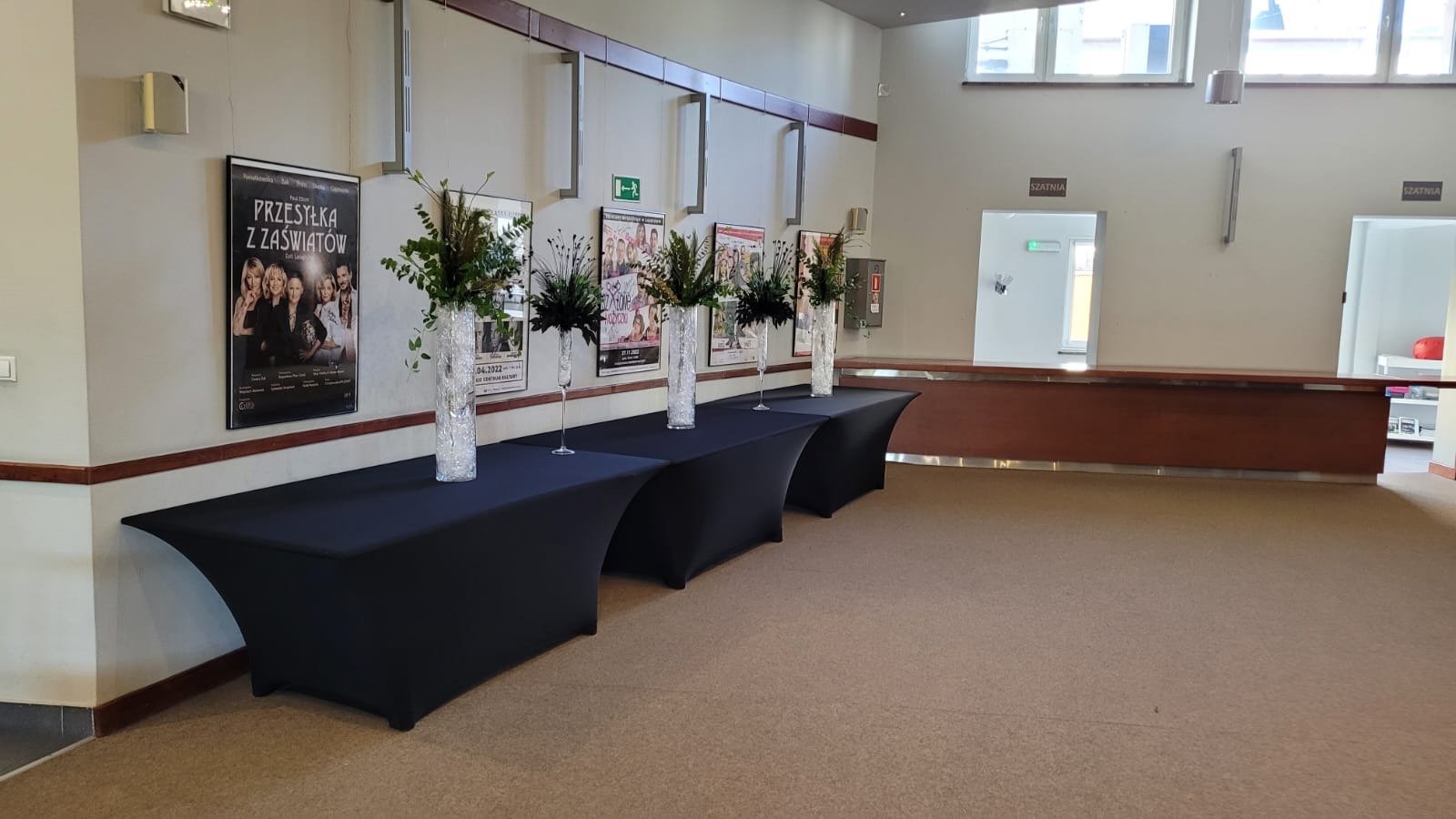 Aranżacja we wnętrzu budynku z czterema czarnymi stołami nakrytymi obrusami, na których stoją wysokie, smukłe wazony z kompozycjami kwiatowymi i zielonymi gałązkami; w tle plakaty na ścianie...