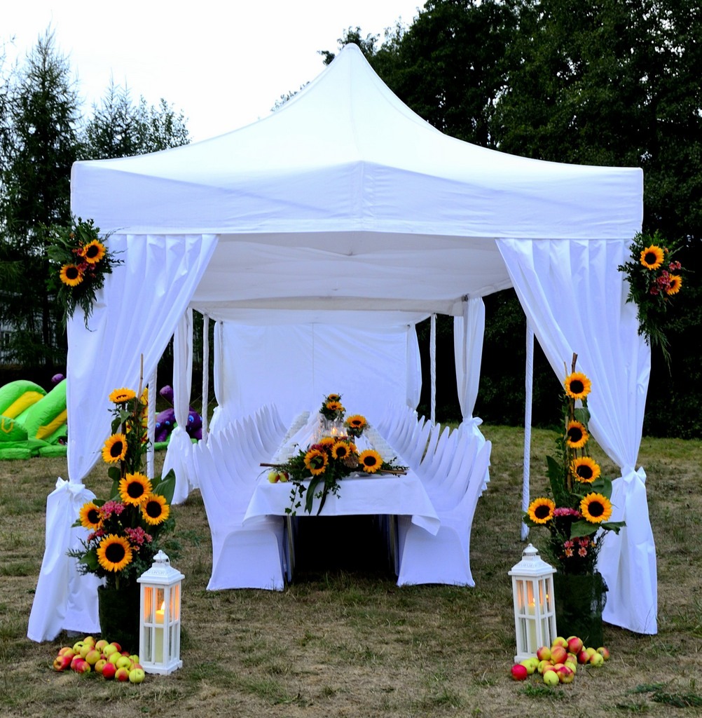 Biały namiot imprezowy udekorowany słonecznikami i jabłkami, stół z białym obrusem i krzesłami w pokrowcach, lampiony ze świecami, w tle dmuchany zamek.
