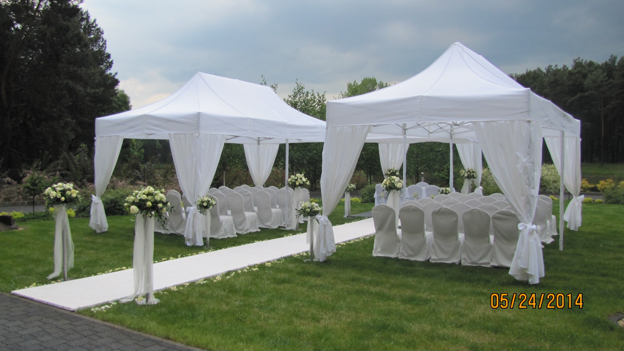 Dwa białe namioty z draperiami i krzesłami, przygotowane na uroczystość na trawniku, dekoracje kwiatowe i dywan w kolorze białym, data 05/24/2014 w prawym dolnym rogu.