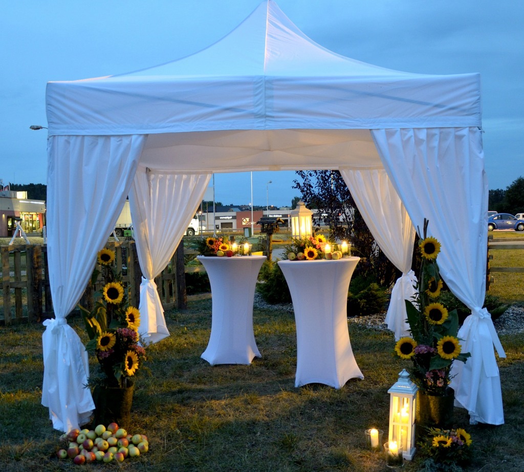 Biały namiot imprezowy z dekoracjami z białych tkanin, słoneczników i jabłek, dwa białe stoliki koktajlowe z kompozycjami kwiatowymi i świecami, lampiony ze świecami na trawie, wieczorne oświetlenie.