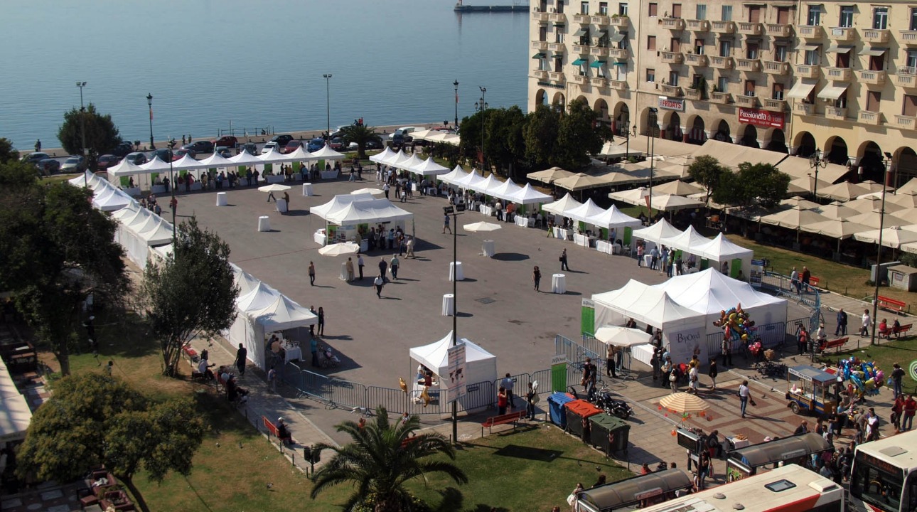 Widok z góry na plac miejski z rozstawionymi białymi namiotami, parasolami i stolikami, prawdopodobnie podczas festynu lub targów, z budynkami w tle i widokiem na morze.