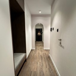 Babka od Wykończeń Magdalena Mazur - Długi korytarz z drewnopodobną podłogą, zabudowaną szafą z siedziskiem po lewej stronie i gładkimi ścianami po prawej, zakończony łukowatym przejściem z czarnymi drzwiami.
