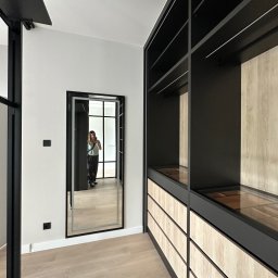 Babka od Wykończeń Magdalena Mazur - Nowoczesna garderoba z czarnymi elementami i drewnianymi frontami szuflad. W lustrze odbija się osoba robiąca zdjęcie. Jasna podłoga i białe ściany.