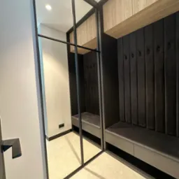 Nowoczesna garderoba w przedpokoju: zabudowa z siedziskiem, wieszakami i szafkami. Lustrzane drzwi przesuwne z czarnymi profilami. Stonowana kolorystyka, eleganckie wykończenie.