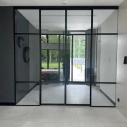 Nowoczesne, czarne, przeszklone drzwi przesuwne oddzielające wnętrze od ogrodu. Widoczne dwa okrągłe zegary na ścianie. Jasna podłoga.